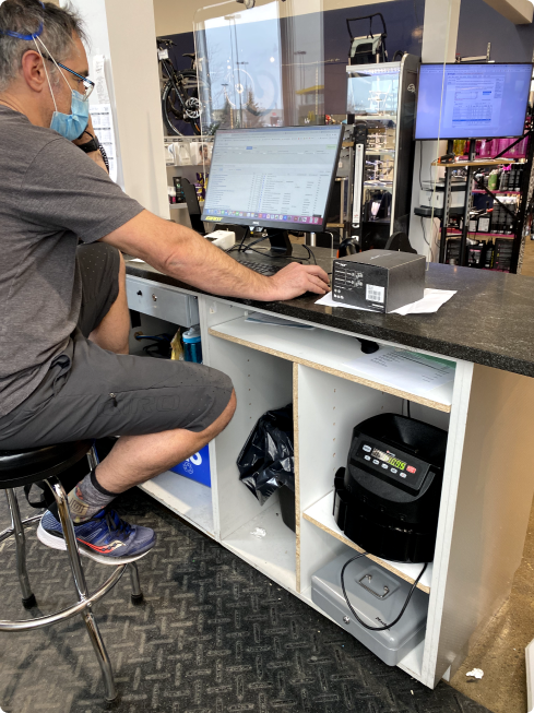 A merchant seated at their store counter using Lightspeed on the POS monitor, with a receipt printer, cash drawer, and shop fixtures around them