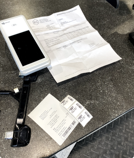 A close-up of the merchant's counter: a payment terminal, a folded bordereau d'expedition packing slip, and a strip of printed barcode shipping labels