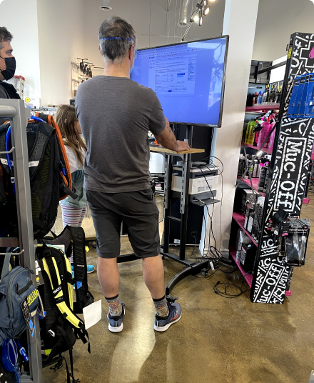 A merchant standing at a floor-level workstation using Supplier Market on a large wall-mounted screen, with backpacks and bike accessories on display and a customer nearby